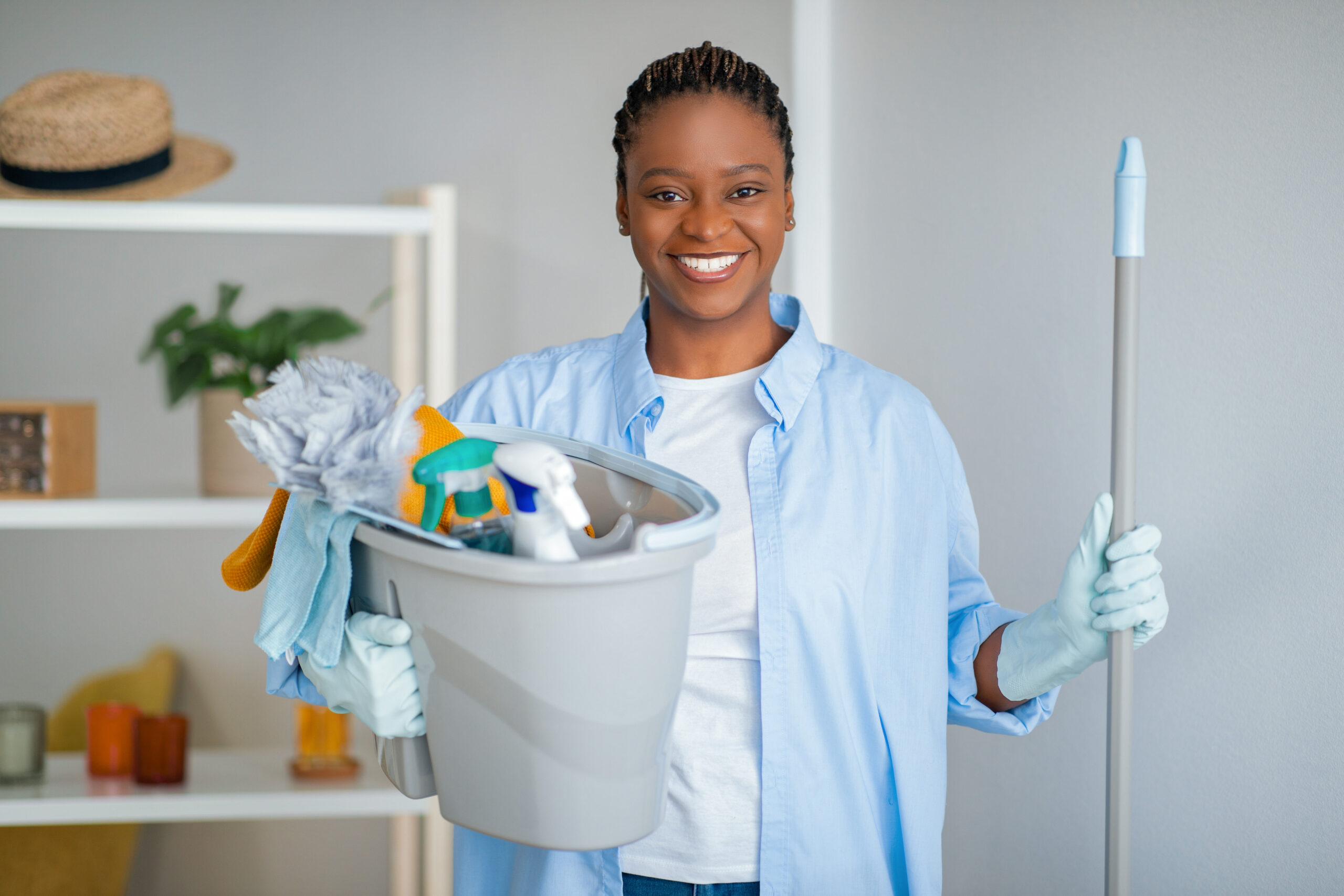 Friendly happy young african american lady in workwear maid ready for cleaning house, black woman holding basket with cleaning tools and mop, smiling at camera. Cleaning service, house-keeping